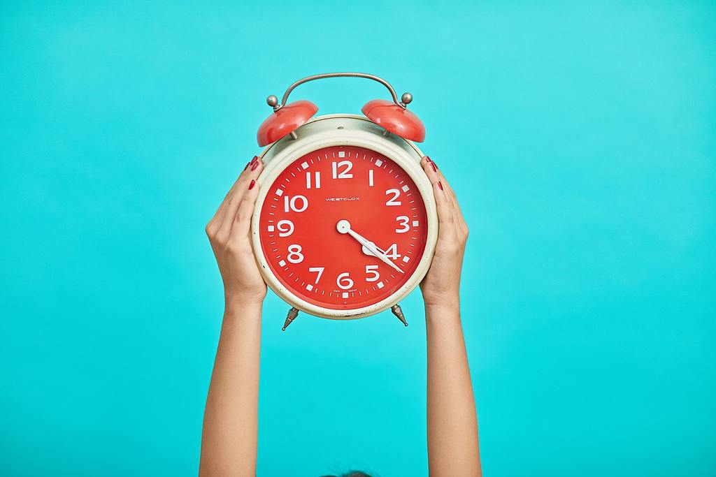 Photo of an orange and white clock held in the air with a hand on both sides on a blue background