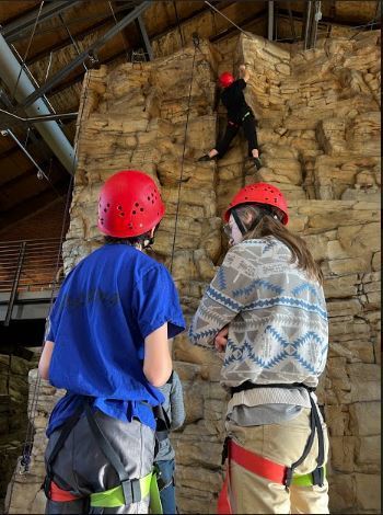 Image of two students in red helmets and climbing gear watching another student  on a climbing course